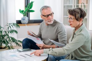 mature couple sit on couch, calculate expenses and planning monthly budget, at home. serious husband and wife filing tax application in living room.